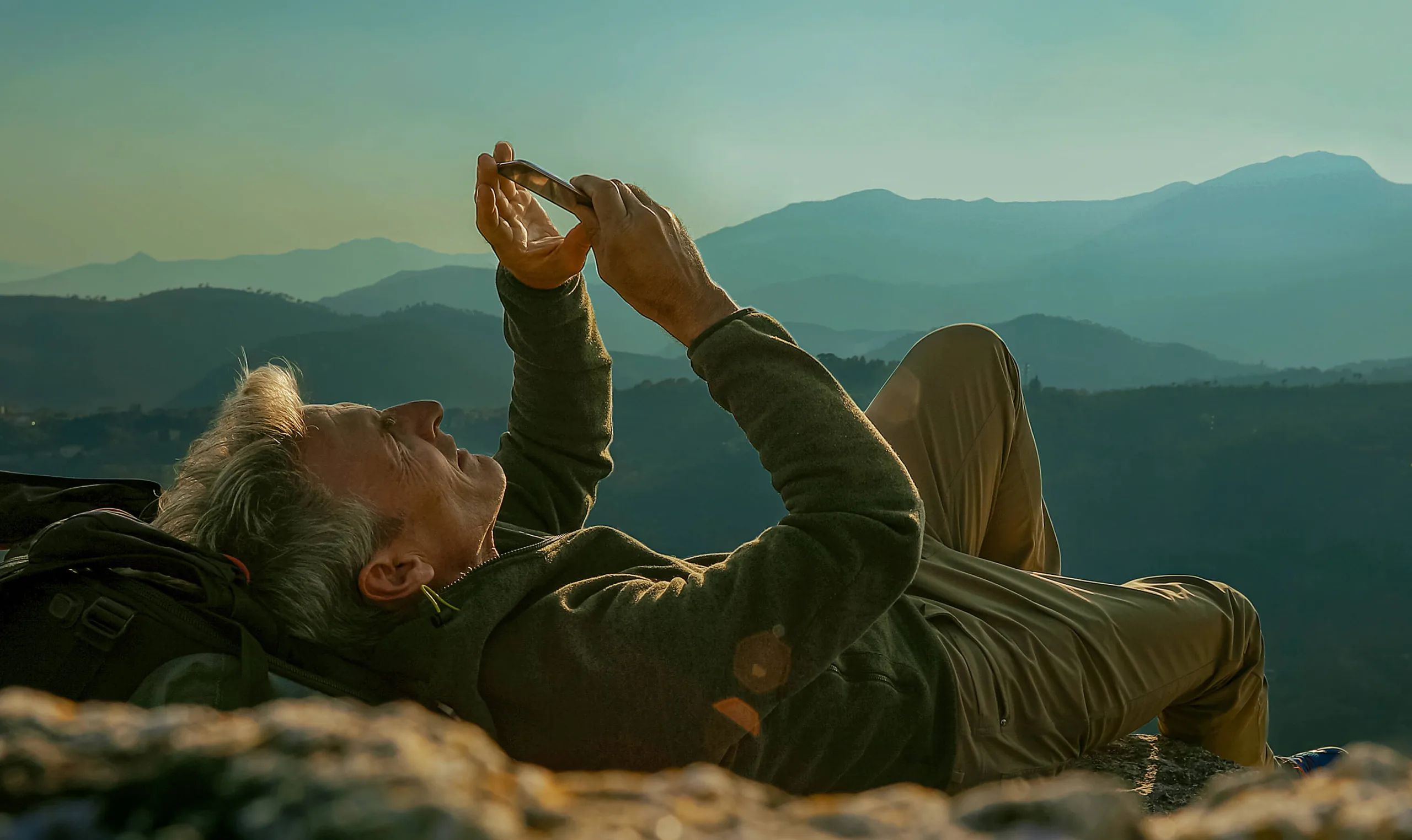 Senior laying down looking at phone while on top of mountain